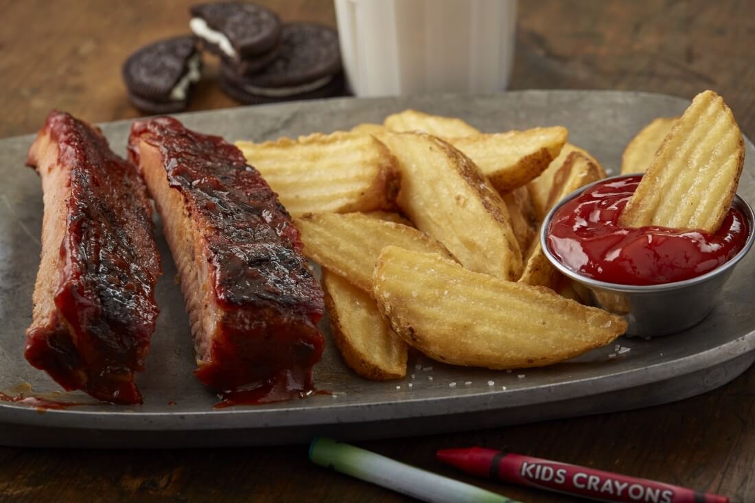 Two individual ribs in BBQ sauce, a few wedge fries, and a small ramekin of ketchup on a gray plate