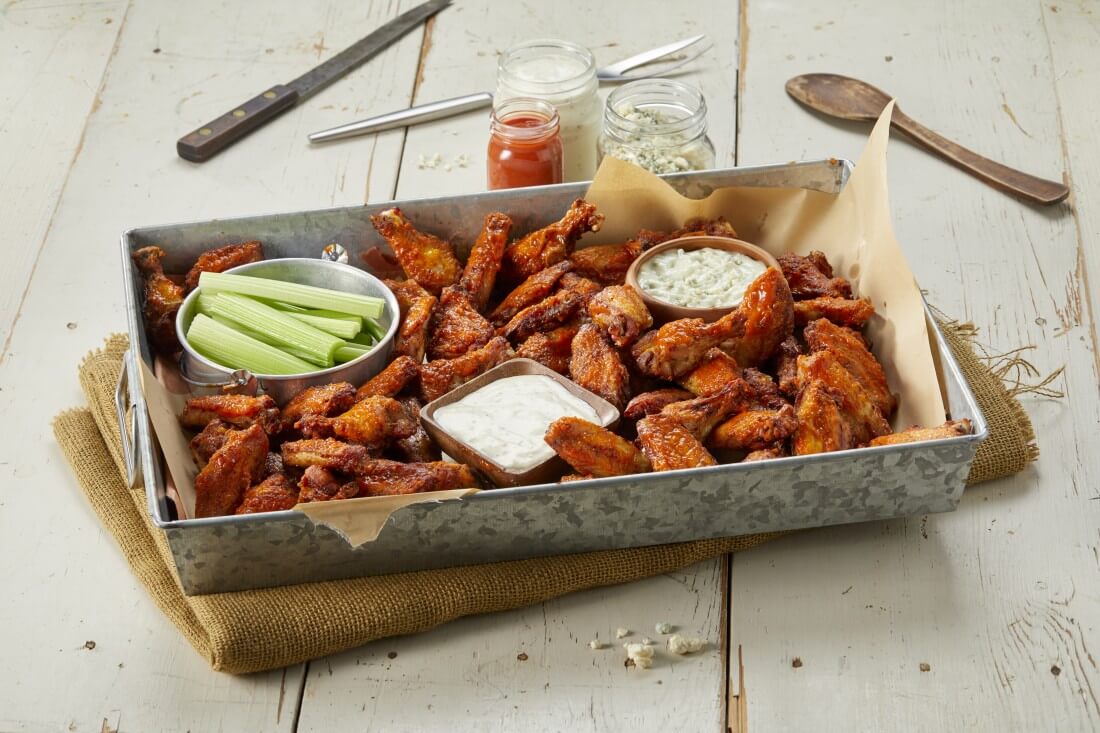 A square, metal tray filled with chicken wings, with a bowl of celery sticks, a bowl of ranch, and a bowl of blue cheese dressing between the piles of wings