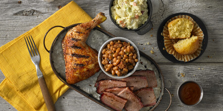 A golden roasted chicken thigh, bowl of baked beans, and sliced meat on a metal tray that sits on top of a yellow napkin, surrounded by a bowl of cole slaw and a cornbread muffin