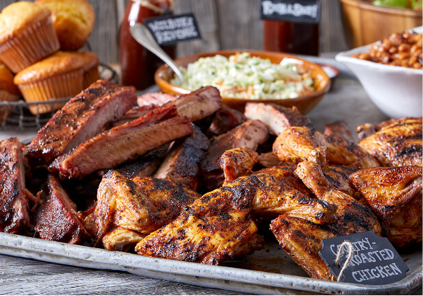 A large platter with Famous Dave's country-roasted chicken and a pile of ribs, with a basket of cornbread muffins, a bowl of cole slaw, and baked beans in the background