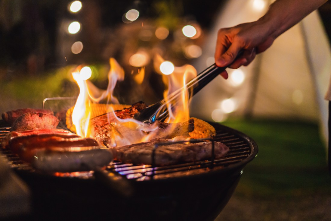A person holding tongs over a grill at night time loaded up with sausages, steaks, corn, and others.