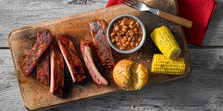 Five ribs cut up on a wooden cutting board, accompanied by a side of baked beans in a white bowl, corn bread, and two piece of corn on the cob