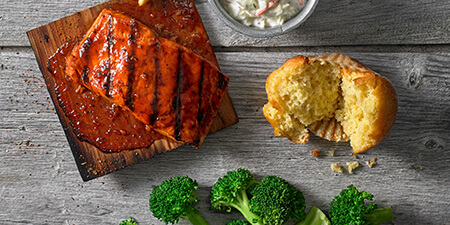 A salmon filet with BBQ sauce on it on a plank of wood, with a cornbread muffin and pieces of broccoli on the side
